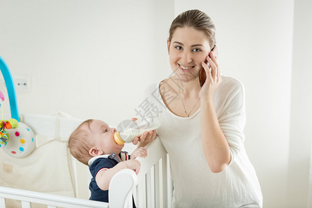 微笑的年轻女子用电话说同时喂养婴图片