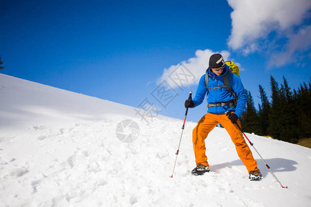 一个背着登山杖和背包的年轻人在蓝天和云彩的映衬下在山上的雪地上行走图片