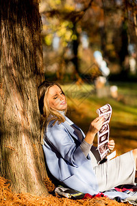 年轻孕妇坐在秋季公园的一棵树下看着背景图片