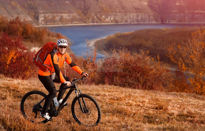 年轻的山地自行车手在河下骑自行车反对美丽的风景穿着橙色运动夹克头盔太阳镜和红色背包的骑自行车者春暖花开的图片