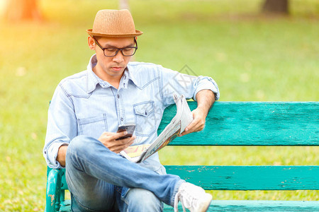 坐在木板凳上戴帽子的亚洲青年男子阅读报纸及智能手机屏幕校验消息图片