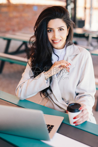 垂直的年轻黑发女企业家享受安息在咖啡馆与电子设备和咖啡检查电子邮件上的笔记本电脑使用的无线连接直视摄像机的肖像图片