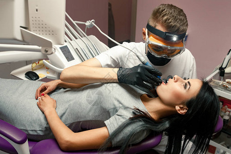 年轻女子在牙科诊所治疗牙齿戴着手套防护眼镜和面罩的男牙医用牙钻图片
