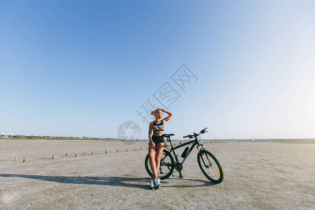 身着五彩西服的金发强壮女人站在沙漠地区的一辆自行车旁图片
