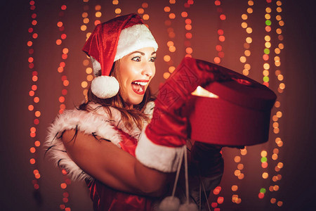 穿着圣诞老人服装的快乐美丽的年轻女子在打开礼物时享受着图片