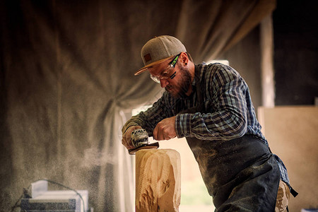 有经验的木工穿着工作服小木匠老板在木工车间工作用角磨机在桌子上加工板是锤子和许多工具图片