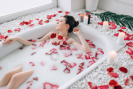 年轻女子躺在浴缸与泡沫玫瑰花瓣和燃烧蜡烛装饰图片
