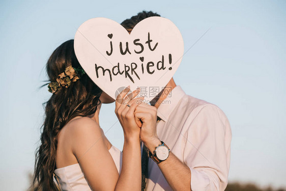 年轻的新婚夫妇亲吻和持有的心与刚刚结婚题词图片