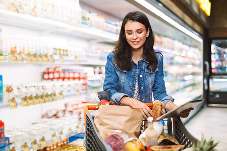 微笑的女孩在牛仔夹克与手推车装满的产品手持片剂在现代超市乳品部的消费时间图片