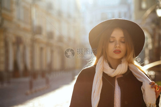 时尚金发女子的户外肖像戴着帽子围巾和大衣摆在阳光刺眼的老街上文本空间图片