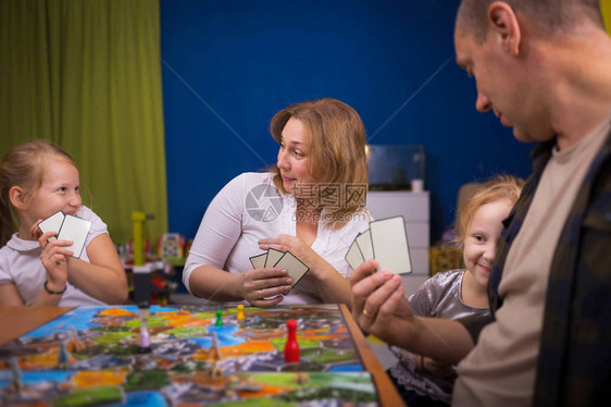 棋盘游戏概念一家人正在家里玩棋盘游戏棋盘游戏场许多数字硬币和沙漏四个人在脸上玩持卡片的快乐情绪图片