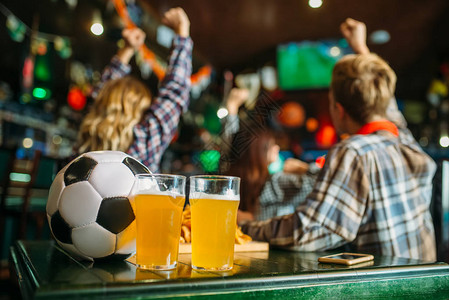 球和啤酒在体育酒吧的桌子上足球迷在后台电视广播观看游戏概念图片