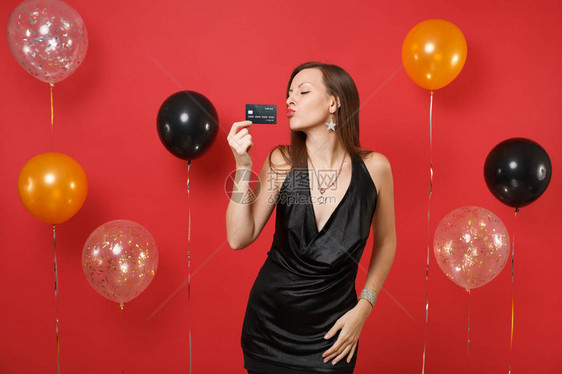 感的年轻女人穿着小黑裙庆祝拿着信用卡吹着嘴唇在鲜红的背景气球上送空气吻新年快乐生日模拟节日聚会理念图片