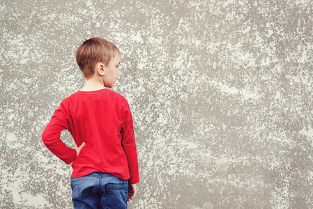 小男孩看墙的背影灰色混凝土墙上的后视图小样穿着红色衬衫和牛仔裤的酷男孩回到学校的概图片