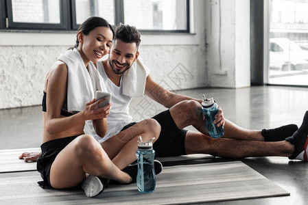 快乐运动的年轻夫妇在瑜伽垫上休息在健身房一起使用智能手机图片