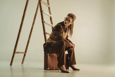 漂亮时尚的女人坐在米黄色梯子附近的老式手提箱里图片
