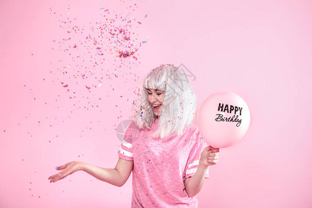 穿着银色头发的粉色t恤的风趣女孩在粉红色的背景上露出微笑和情绪有气球的少妇或女孩生日快乐从上面扔五彩纸屑假期背景图片