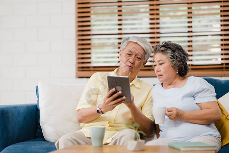 亚洲老年夫妇在家中使用平板电脑在客厅喝咖啡在家休息时躺在沙发上享受爱情时刻享受时间生活方式老年家庭在家的概念图片