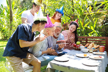 快乐的多民族家庭在高加索人爷爷生日快乐之际给他送上惊喜礼物阳光明媚的日子他在后院的户外吹灭自制蛋糕上的蜡烛图片