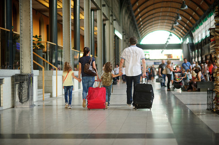 年轻的父母带着两个孩子在车站一家人穿过一个大等候室都牵手爸妈带着大手提箱背着包的男图片