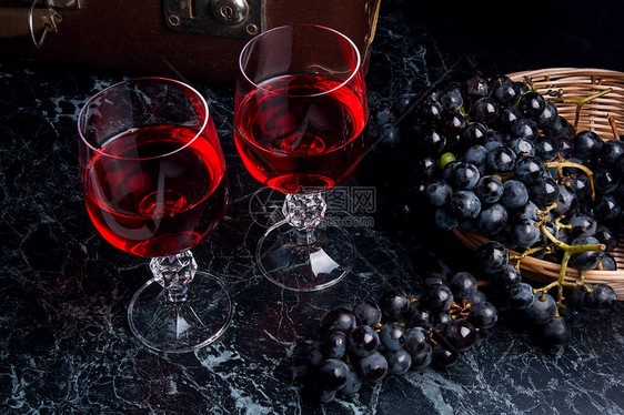 两杯红酒在深色大理石背景上深色大理石背景上的蓝色成熟葡萄黄色篮子里的成熟葡萄和背图片