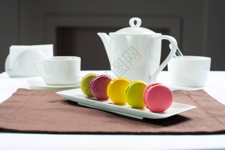白色方形盘子上的彩色马卡龙甜马卡龙和鲜花以白色茶具为背景图片