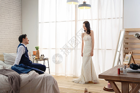 年轻丈夫坐在床上欣赏妻子穿婚纱的样子图片