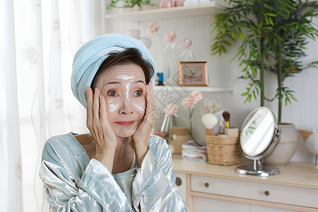 衰老老年女性正在使用面膜头上裹着毛巾坐在桌前对着镜子背景