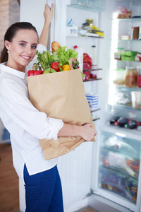 年轻女子捧着蔬菜杂货店购物袋。站在厨房里。在厨房里看着相机的女人