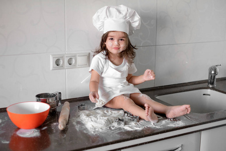 可爱的小女孩在围裙和厨师的帽子是揉面团和烘烤，同时微笑着