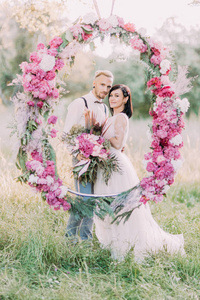 新婚夫妇手持牡丹花束, 站在阳光明媚的春天田野里的牡丹拱门后面。特写垂直照片