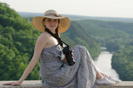 戴着帽子的女孩坐在山与相机上的森林和蜿蜒的河流背景
