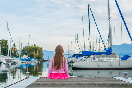 可爱小女孩看着船在湖上，坐在码头上查看从背面