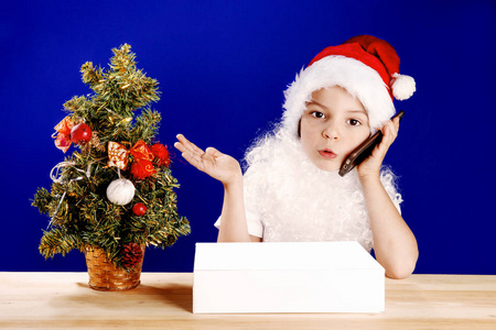小圣诞老人感情谈一部智能手机。在一个白色的盒子在桌子上的礼物。蓝色背景。特写