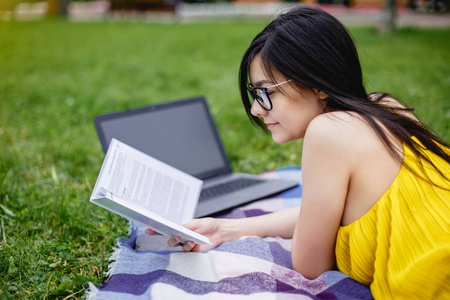美丽的学生女孩读过的书和笔记本计算机在夏季里公园的绿色草地上
