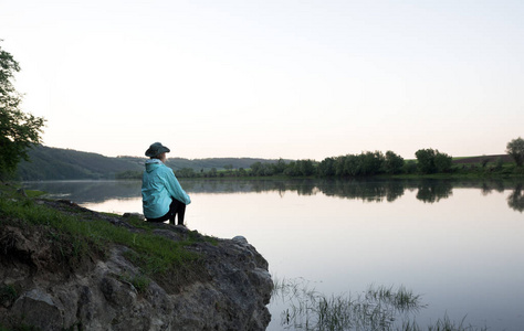 女性旅行者坐在石头上面这条河。日落的时候。小山和森林对银行