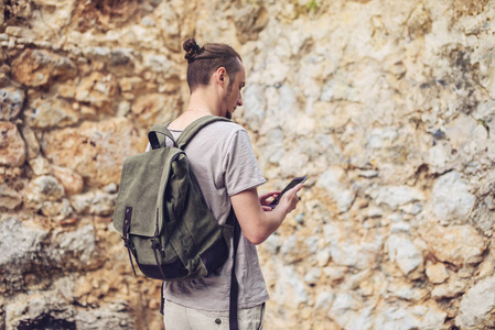 带背包的男子旅行者在石墙的背景下看着他的智能手机