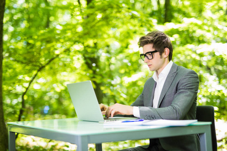 工作的笔记本电脑在绿色公园办公室桌的英俊的年轻商业人的肖像。经营理念