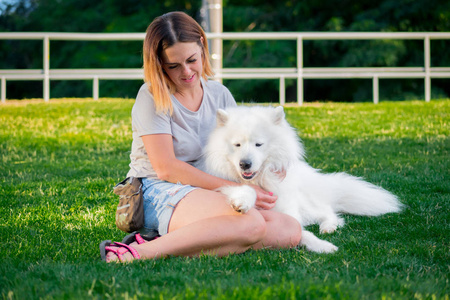 成年红头发女人扮演和抚摸她的萨摩耶德犬的狗。白色蓬松宠物公园里的情妇在绿色的草坪上玩得开心