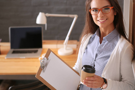 年轻的女子站在桌子旁边与笔记本电脑拿着文件夹和杯咖啡。工作场所。女商人