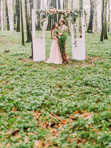 这对新婚夫妇身着复古服装, 站在拱门下, 用红白玫瑰装饰的白色门的形式, 竖着照片。拱门放在绿色的森林里