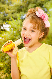 年轻漂亮的女孩，身穿黄色 t 恤，吃健康的香蕉在花园的背景