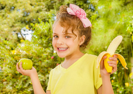 美丽的年轻女孩，穿着黄色的 t 恤，持有健康苹果和香蕉，在花园的背景