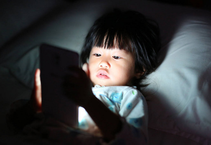 孩子玩智能手机在晚上躺在床上的女孩
