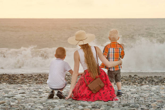 妈妈和两个儿子坐在海滩上注视着海浪查看从背面
