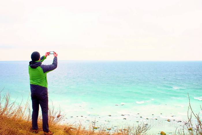 绿色防水夹克的男人需要照片的 coastliny 智能手机。寒冷的大风天海上