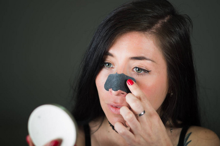 美年轻女子看着镜子的应用黑鼻子面具来清洁皮肤从黑点