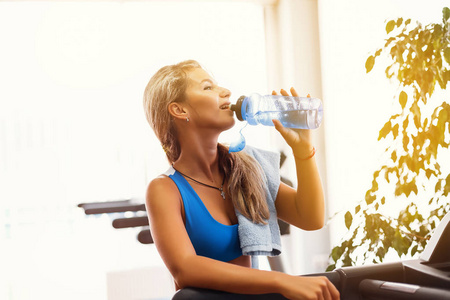 体育金发美女是饮用水在健身房跑步机上