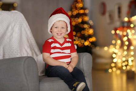 可爱的小男孩，在圣诞老人的帽子，坐在扶手椅上对模糊的圣诞灯
