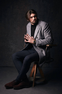 美丽的头发和胡子和严肃的面孔，坐在木椅上，穿着灰色夹克，西服在暗灰色工作室英俊年轻男性模型